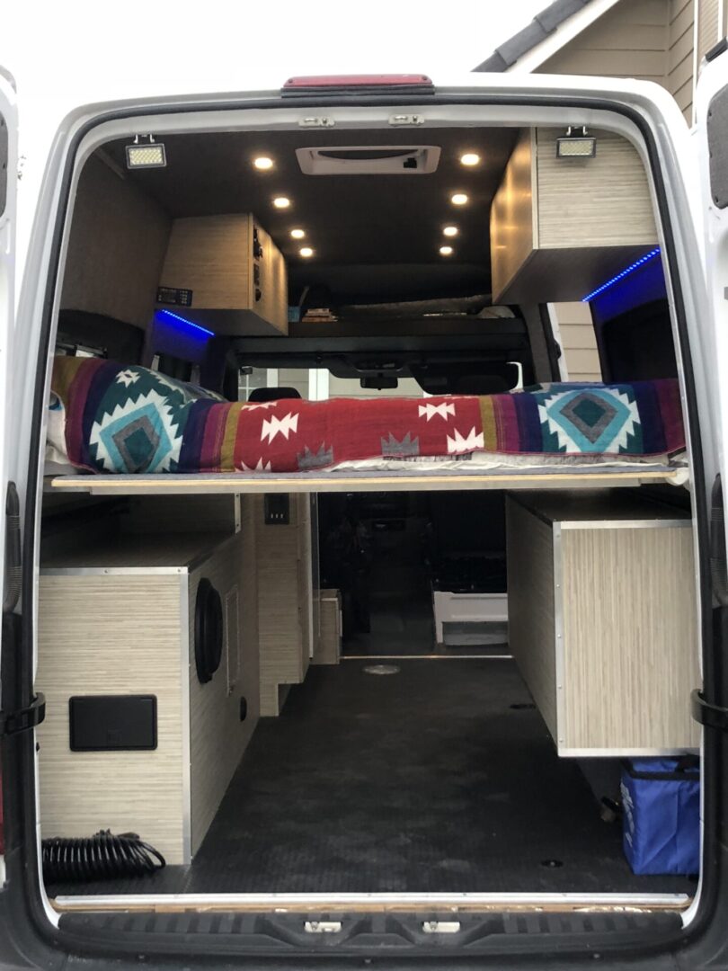 View of a camper van interior with a colorful blanket on a raised bed, wooden cabinetry, and blue LED lights.