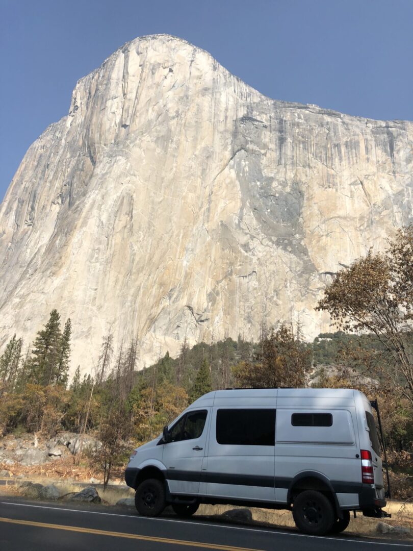 A white van is parked on a road in front of a large rocky cliff in a natural setting.