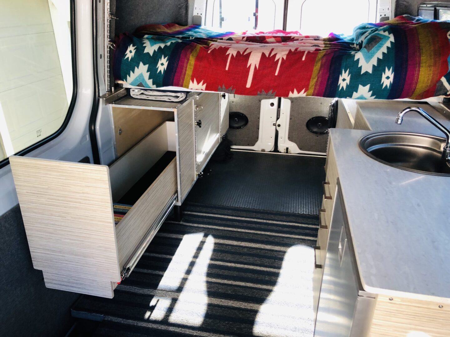 Interior of a camper van with a colorful bedspread, open drawers, a small counter, sink, and striped floor mat.