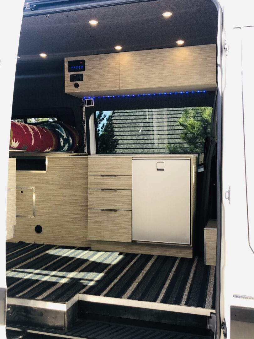Interior of a van with wooden cabinets, striped flooring, and recessed lighting. There's a small bed at the back and a control panel above the cabinets.