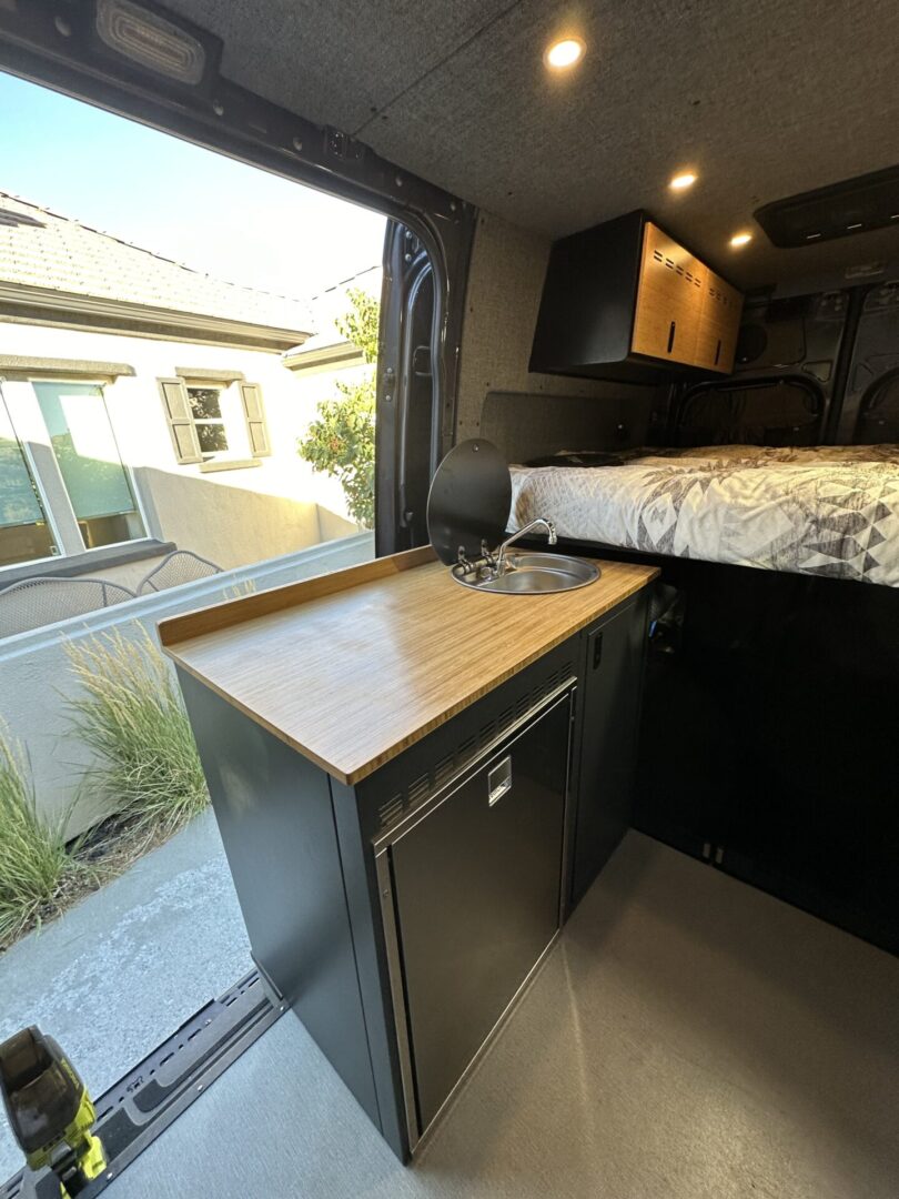 Interior of a camper van showing a kitchenette with a wooden countertop, sink, refrigerator, and overhead cabinets. A bed is visible in the background. Door is open to the outside.