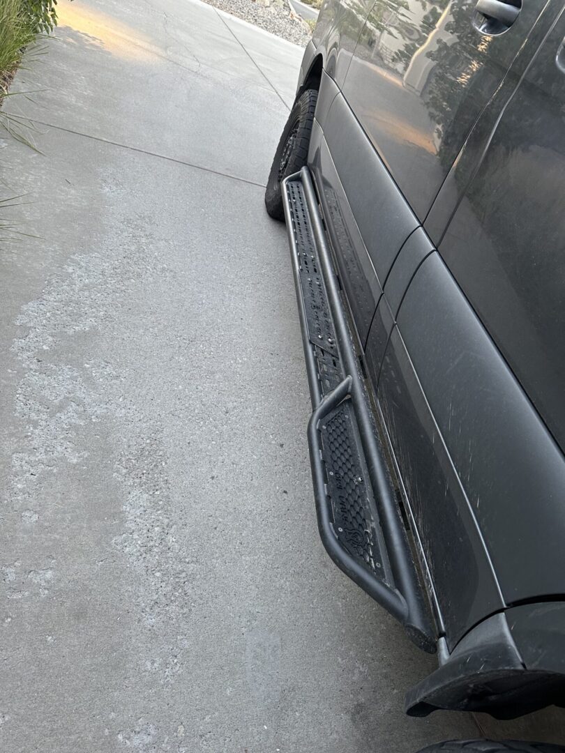 Side view of a gray vehicle on a concrete driveway, highlighting a black step bar beneath the door.