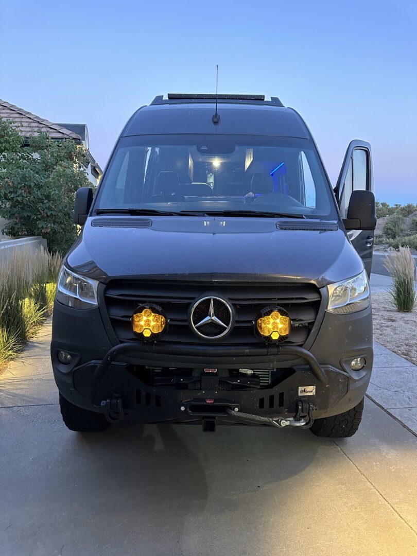 Front view of a dark gray Mercedes-Benz van with illuminated yellow lights and an open driver's door, parked on a driveway at dusk.