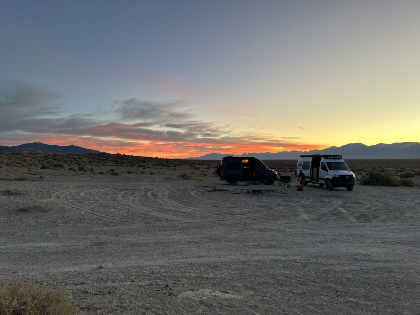 Two vans parked on a barren desert landscape at sunset, with scattered bushes and mountains in the background.