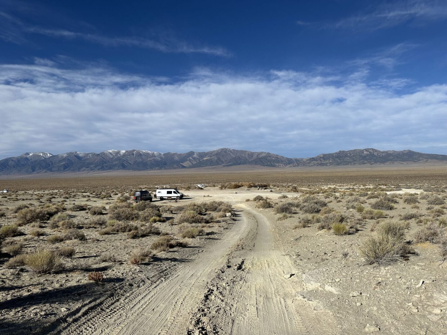 A dirt road leads through a dry, shrub-filled landscape toward parked vehicles near distant snow-capped mountains under a partly cloudy sky.